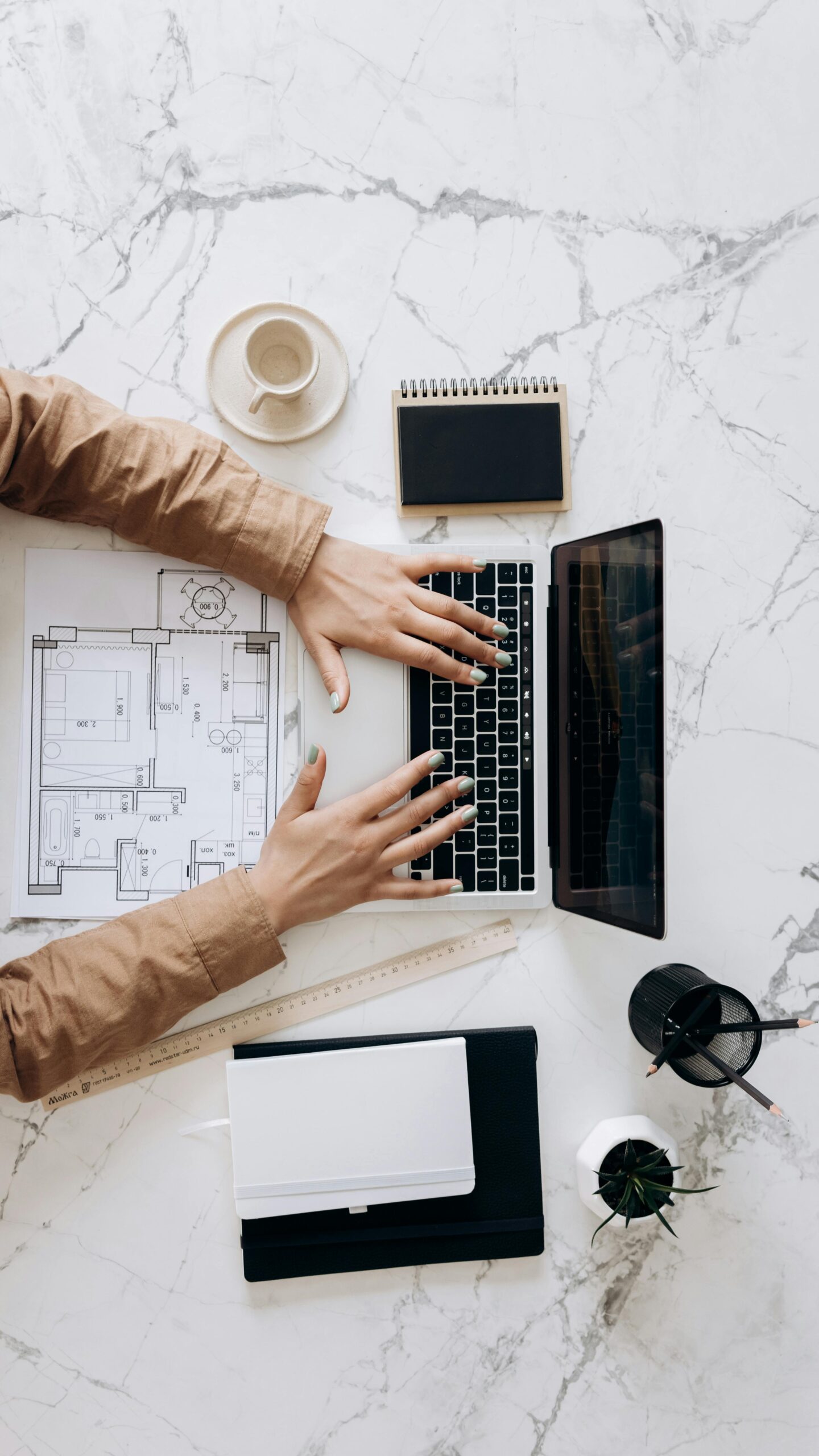 Overhead view of architect using laptop with blueprints and coffee on marble desk.